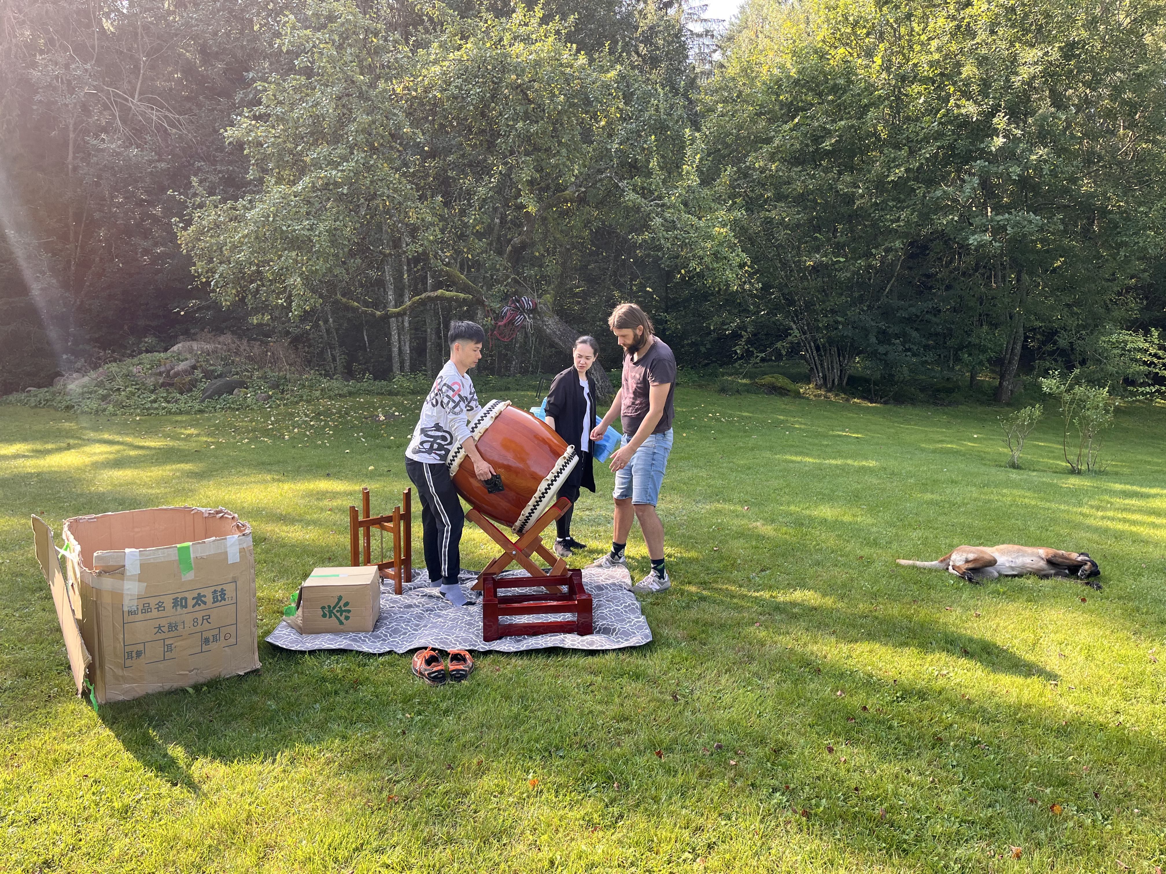 Aki, Augis and Ayano set up a taiko drum on a stand outside on a farm by the woods while Arija the dog lies in the sun 